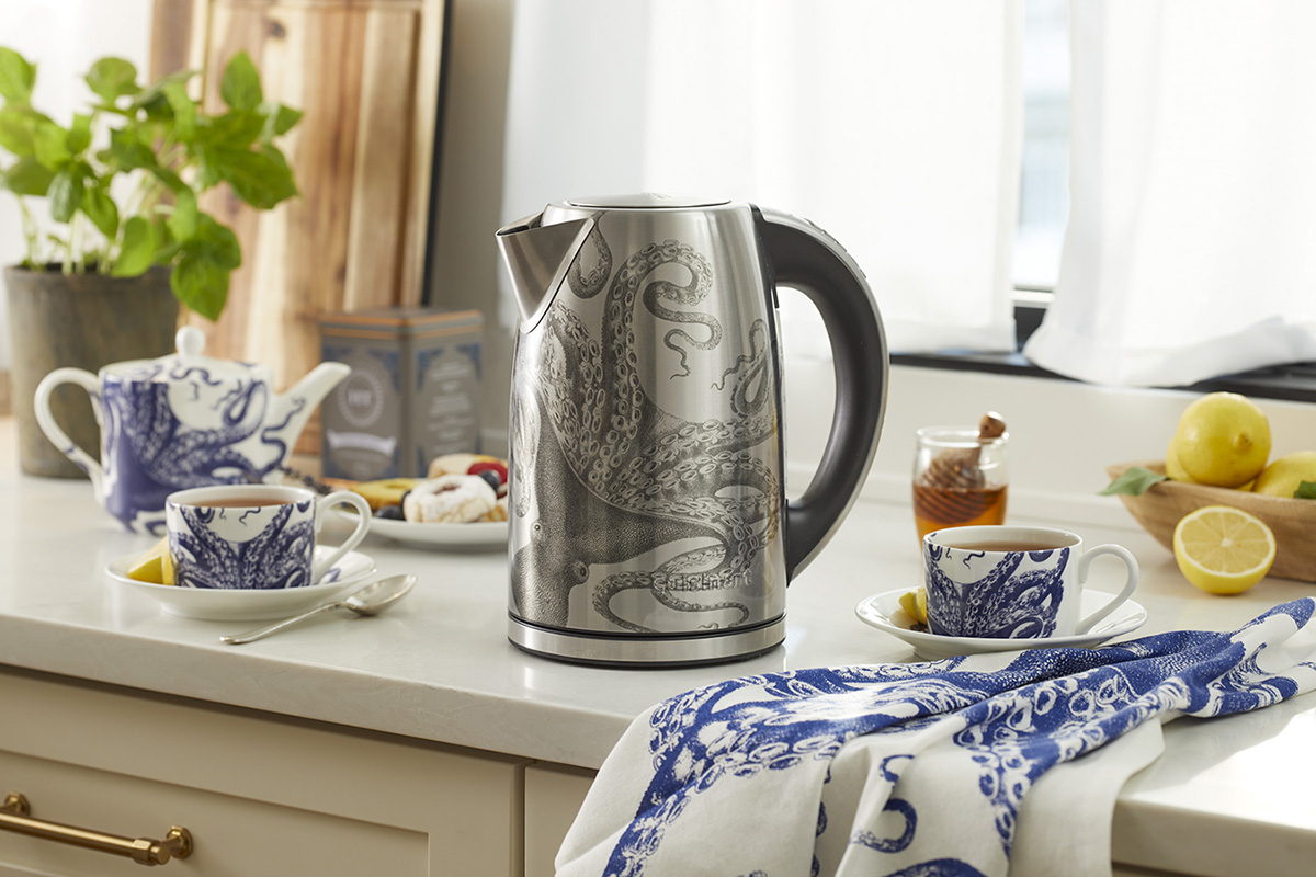 A stainless steel cordless electric kettle with an octopus design on it sitting on top of a kitchen counter next to snacks, a bowl of lemons, teacups, and saucers.