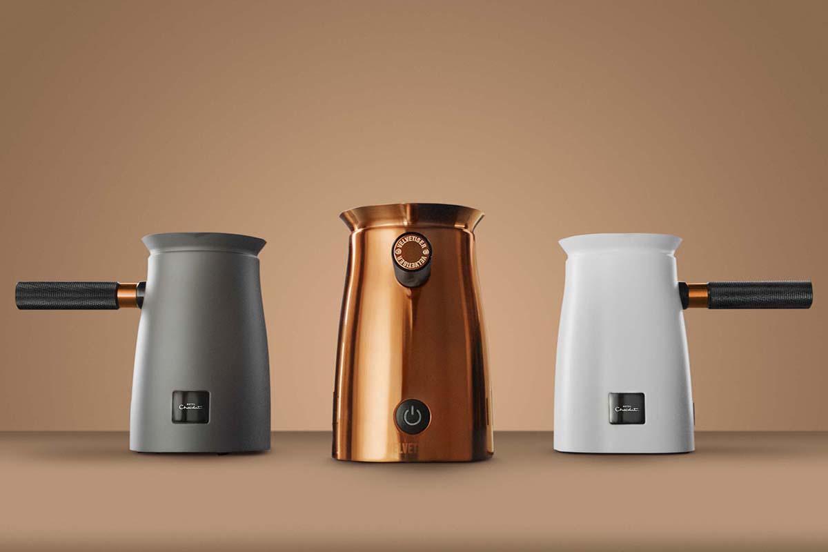 Three gourmet hot chocolate machines in gray, copper, and white that have a chic, stylish and modern design sit on a light brown background.