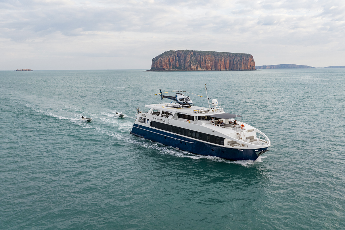 True North expedition cruise line explore Australia's Great Barrier Reef and Whitsundays. A helicopter is landing on top of the yacht while 2 small boats are following it.