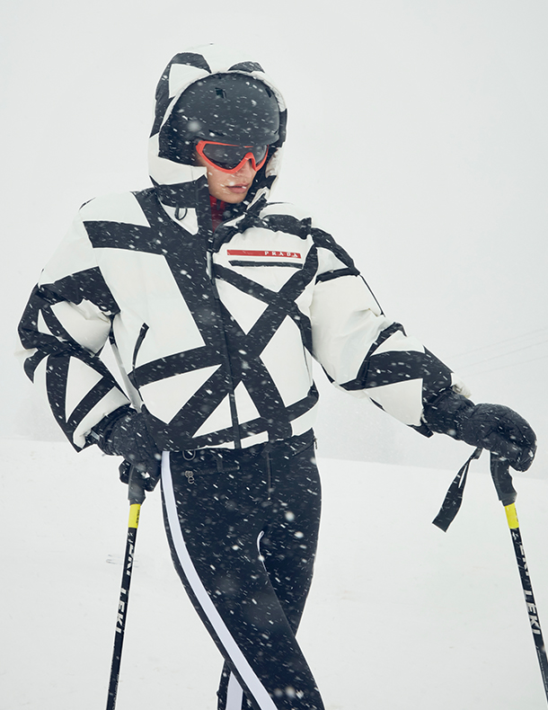 Woman wearing black and white ski jacket with abstract pattern, black ski pants, and sunglasses standing with skis on a snowy background