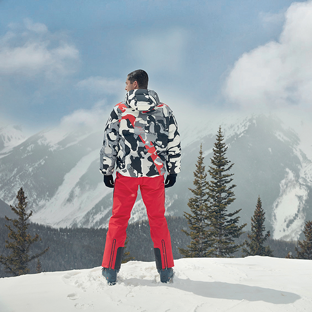 Man dressed in graphic down jacket and red ski pants overlooking snow covered mountains