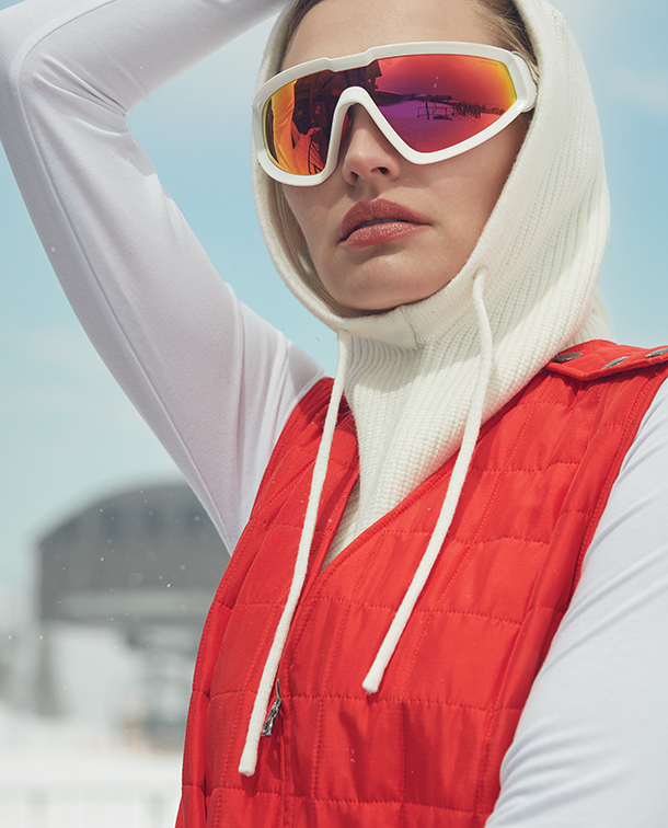 Woman wearing white turtleneck, red ski overall, and white angular shield sunglasses with mirror lenses