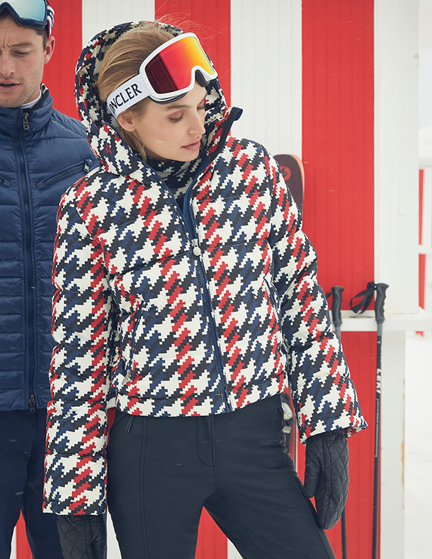 Man in the background wearing navy down jacket and woman in the foreground wearing hooded, down jacket in houndstooth, and white ski goggles