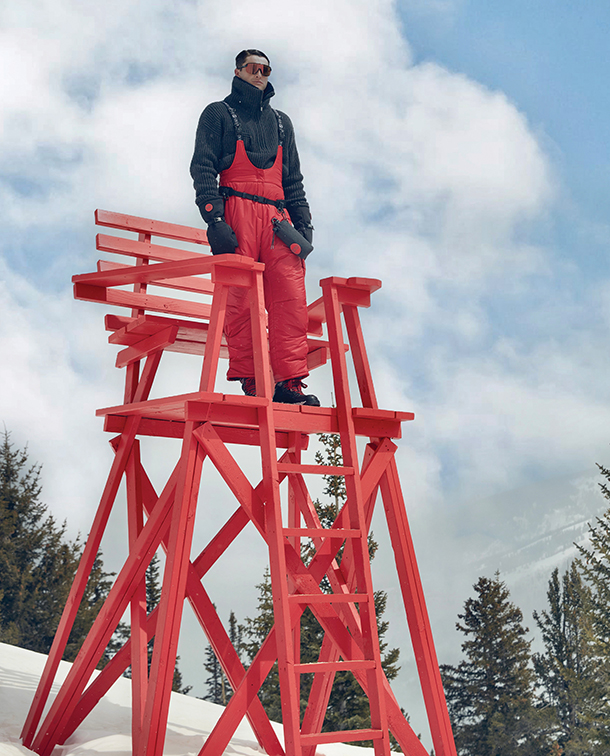 Man dressed in red overalls, black sweater, and sunglasses standing on oversized red wooden chair