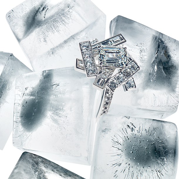 A graphic composition of diamond-set threads frames an emerald cut stone in a sleek and striking diamond ring. The diamond ring is placed between sparkling ice cubes on a white background.