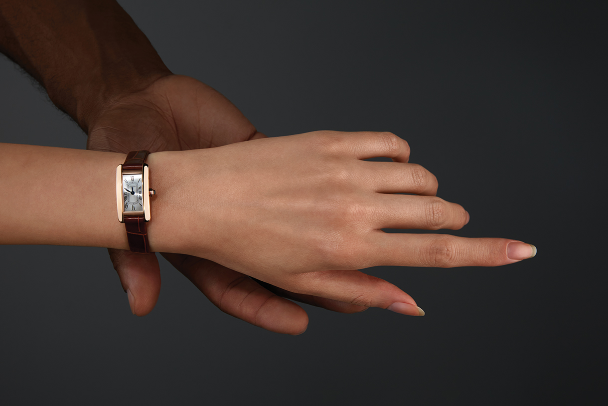 A man’s hand touching a woman’s hand. The woman’s hand has a mini, rectangular-faced watch with gold case, roman numerals, and brown alligator-skin strap.