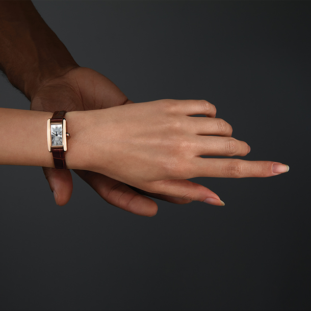 A man’s hand touching a woman’s hand. The woman’s hand has a mini, rectangular-faced watch with gold case, roman numerals, and brown alligator-skin strap.