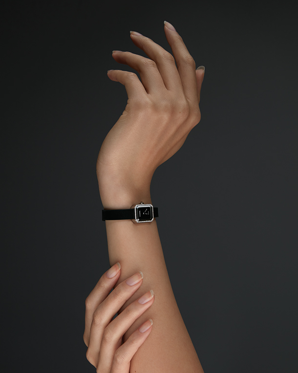 Woman holding her forearm wearing a mini black watch with black face, diamond case, and thin black strap.