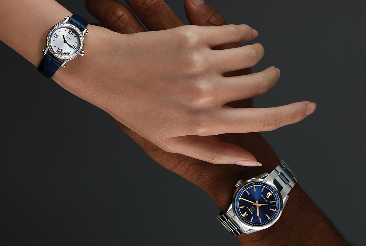 Woman’s hand in front of man’s hand, both wearing watches. The woman’s watch has a diamond bezel, pearl face, and blue leather strap. The man’s watch has a blue dial with gold hands and steel band.