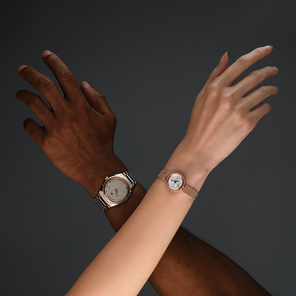 Man and woman crossing arms both wearing slightly smaller vintage-style watches. The man is wearing a steel, rose gold, and sand grey watch. The woman is wearing a gold watch with pearl face.