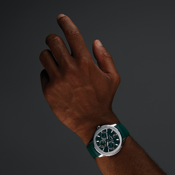 A man’s hand with watch on his wrist. The watch has a green dial, steel case, a perpetual mechanical calendar, and interchangeable green rubber strap.