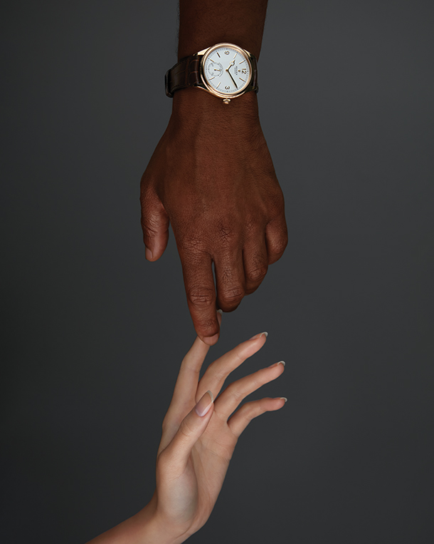 Woman and man touching forefingers. The man is wearing a watch with a round white dial, yellow gold case, and brown strap on his wrist.