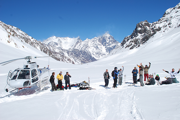 A heli-ski helicopter parked on a snow-capped mountain with ski guides and several skiers waiting next to it.
