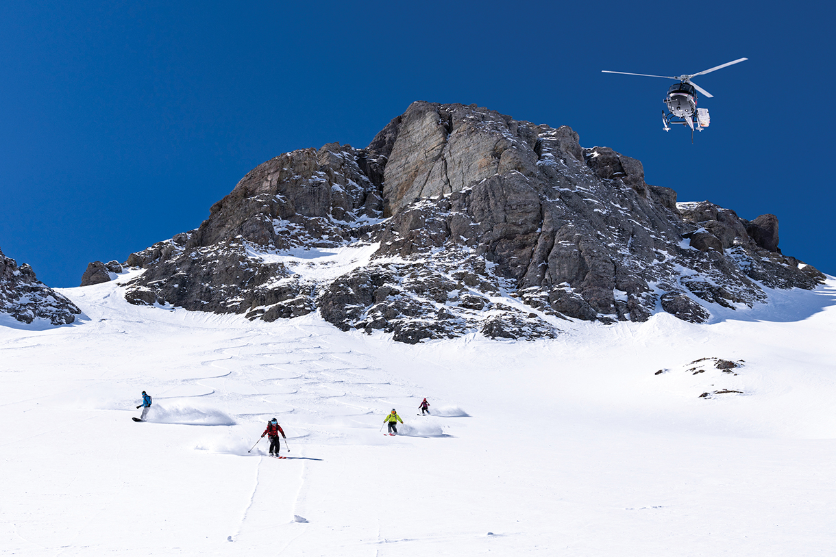 A helicopter flying over a snow-capped mountain with four skiers skiing below.