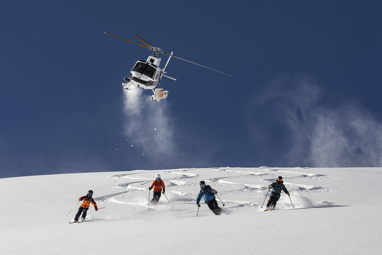 A heli-ski helicopter is flying over a snow-covered mountain while skiers are skiing downhill right below.