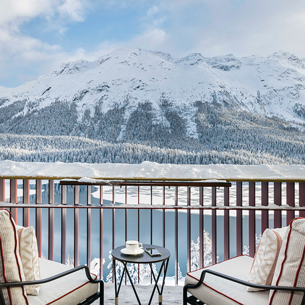 A hotel balcony with two chairs and a table overlooking snow-capped mountains and a frozen lake.