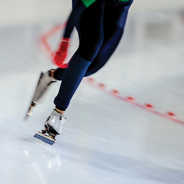 Close up of a speed skater wearing a dark blue skin suit, red gloves and white speed skates gliding on an indoor ice rink.