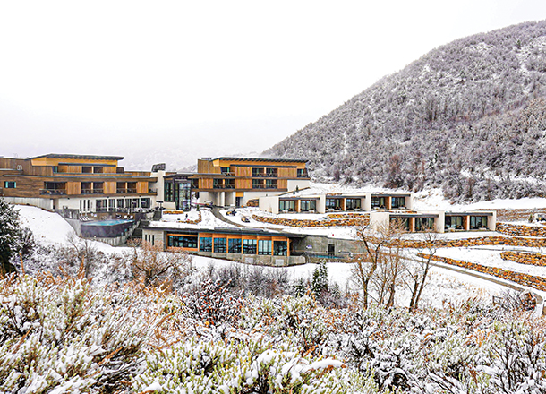 A group of wooden chalets at an alpine resort on a white snow-covered hillside. The buildings are surrounded by snow-covered trees and mountains.
