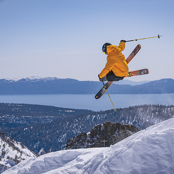 A skier in a yellow orange ski jacket and black pants doing a jump down a snow-covered mountain slope. There are mountains and a lake in the background.