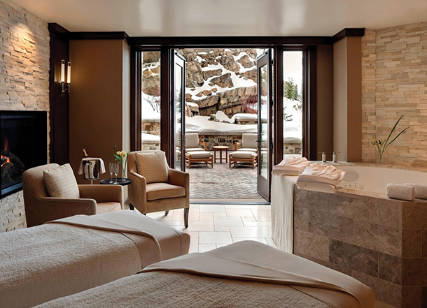 A spa treatment room at a resort with two beds, a hot tub, large floor-to-ceiling glass doors, a fireplace and an outdoor patio with seating outside.