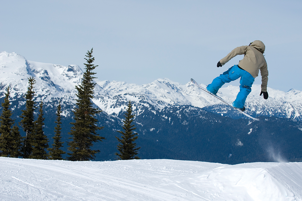 A snowboarder wearing a brown snow jacket and blue pants is doing a jump over a snow-covered slope. There are tress and snow-covered mountains in the background.