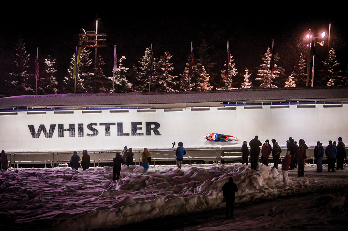 Skeleton sledding at night at the Whistler Sliding Center with a people standing and watching.