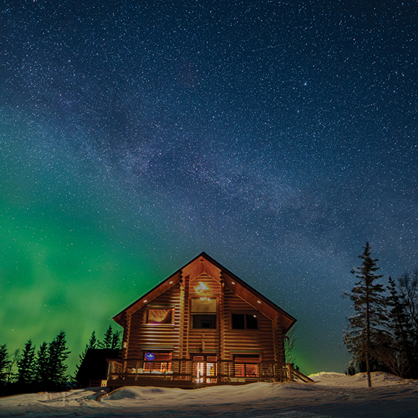 A log cabin in the snow under a colorful starry night sky.