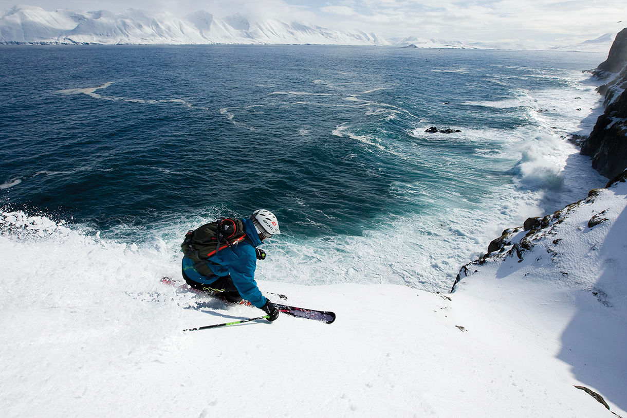 A heli-skier skiing a run from snowy summit to sea in North Iceland. Below is a rocky cliff and the artic sea