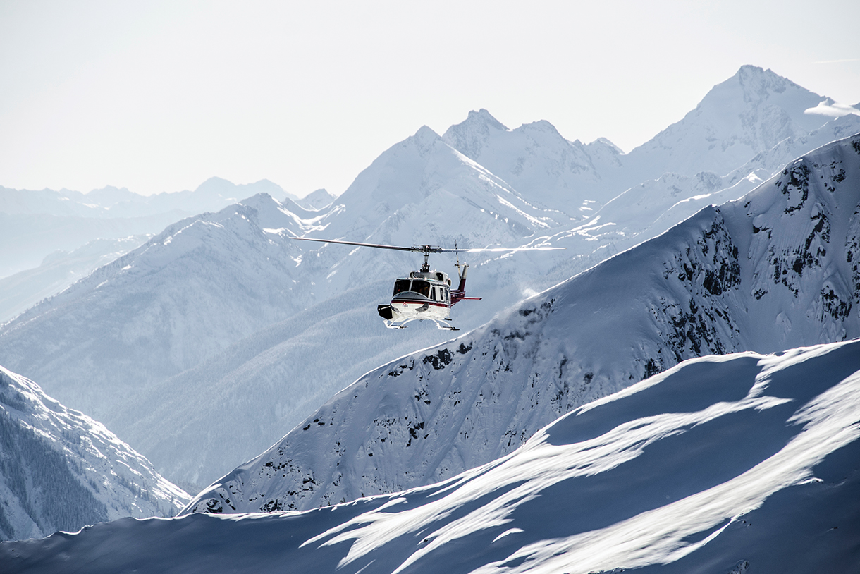 A heli-ski helicopter is flying over a snow-covered mountain range.  Caption: Heli-skiing in British Columbia, Canada