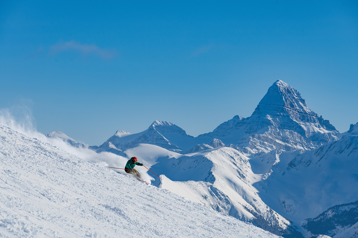 A person skiing down a steep snow-covered mountain slope. In the background there are snow covered mountain peaks.