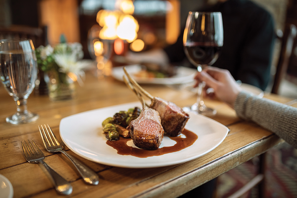 Close up of rack of lamb entree, served on a white plate in a restaurant. The table setting is on a wooden table. A person is holding a wine glass with red wine.
