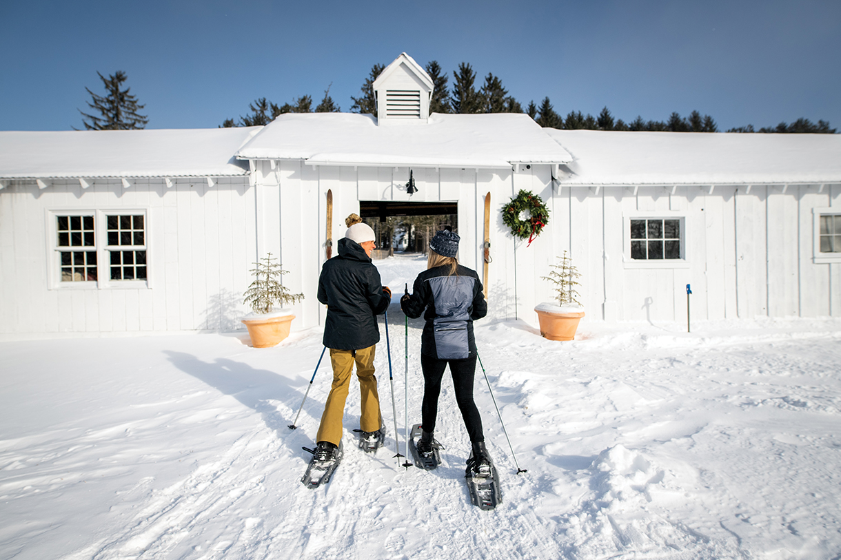 A couple with skis walking on snow toward Edson Hill’s Nordic Center, a white historic barn-styled property for winter activities.