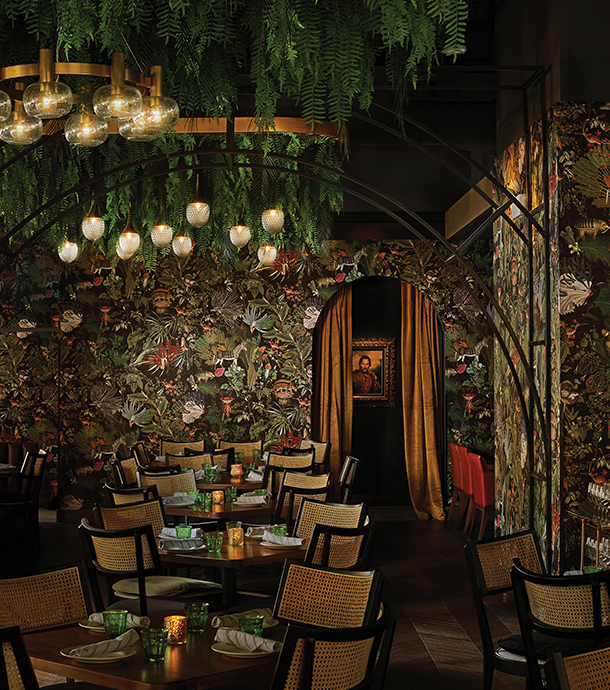Interior of rustic restaurant with several tables and chairs set up for dining. There are chandeliers hanging from greenery and wall paper designed with foliage.