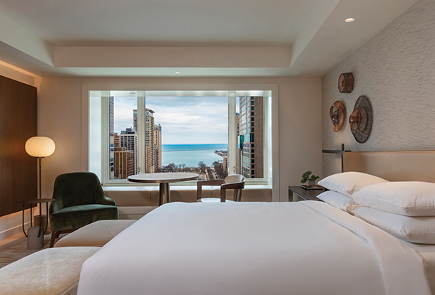 A hotel room decorated in neutral colors that has a king-size bed with white bedding. There is a small table with chair next to a large window which reveals a view of the Chicago’s city skyline. 