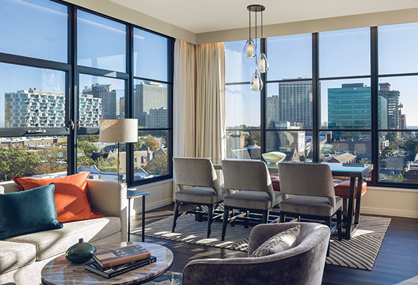 A large hotel room featuring a living and dining room space with a view of a city skyline. The décor is modern and sophisticated. The windows are dramatic with floor-to-ceiling views. 