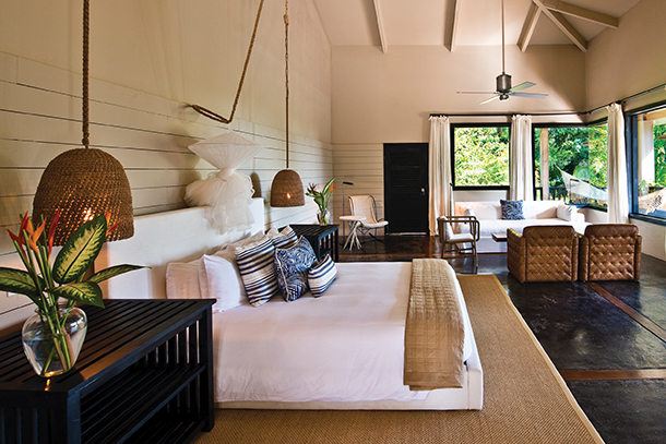 A guest room with a king-size bed, a couch, chairs, and a ceiling fan in neutral colors. The room has a vaulted ceiling with exposed wooden beams and large windows with white curtains. 