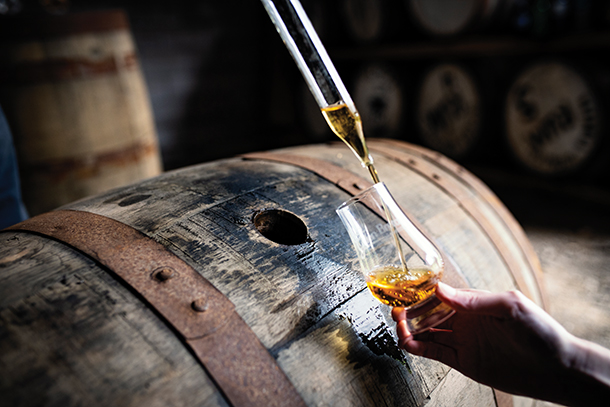 A person pouring whiskey into a glass from a wooden barrel. The barrel is large and appears to be in a dimly lit warehouse. 