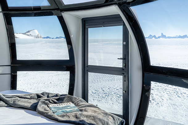Futuristic, semi-ensuite bubble accommodations at Echo Camp. Closeup of curved, floor-to-ceiling glass panels with views of white icy landscape. There is a bed with a blanket and book on top of it.