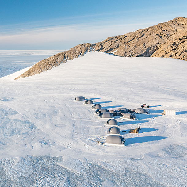 Gray, space-inspired, bubble accommodation pods at a remote base camp in Antarctica’s pristine white interior mountain range. Brown pitted rock formations protrude from Henriksen Nunataks’ glaciers.)