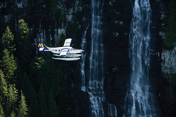 A small white seaplane flying over a cascading waterfall which is surrounded by green trees. The seaplane has two pontoons and has the company logo on its tail.  