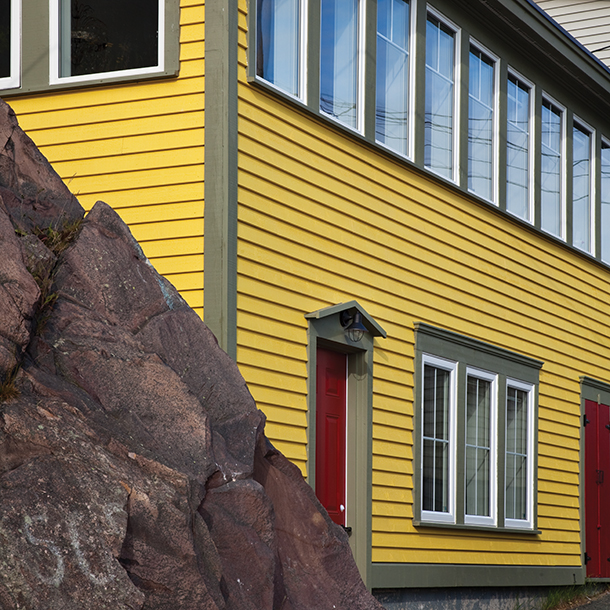 A yellow house with many windows next to a large rock. The house is two stories tall with gray and white trimmed windows and red doors.