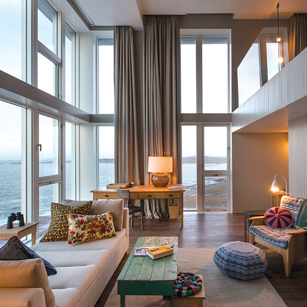 A cozy living room with large floor-to-ceiling windows offers a spectacular view of the ocean. The room is decorated in a modern Scandinavian style, with light walls, wood floors, and pops of color from the artwork and furniture. 
