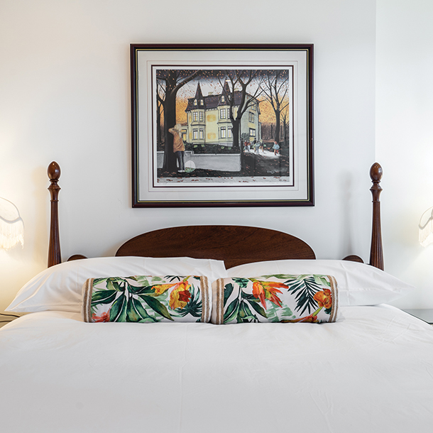 A bedroom with a bed and a colorful landscape painting hanging above the bed. The bed has a dark wood frame and white bedding. There are two white pillows and two colorful patterned pillows on the bed. 