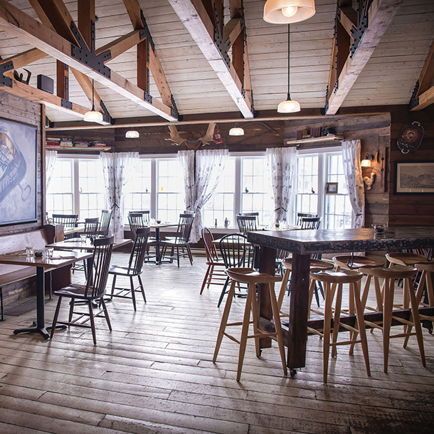 A restaurant dining room with wooden tables and chairs, decorated in a rustic style with exposed beams and stone walls. There is artwork displayed on a wall and large windows at the back of the room which offer scenic views. 