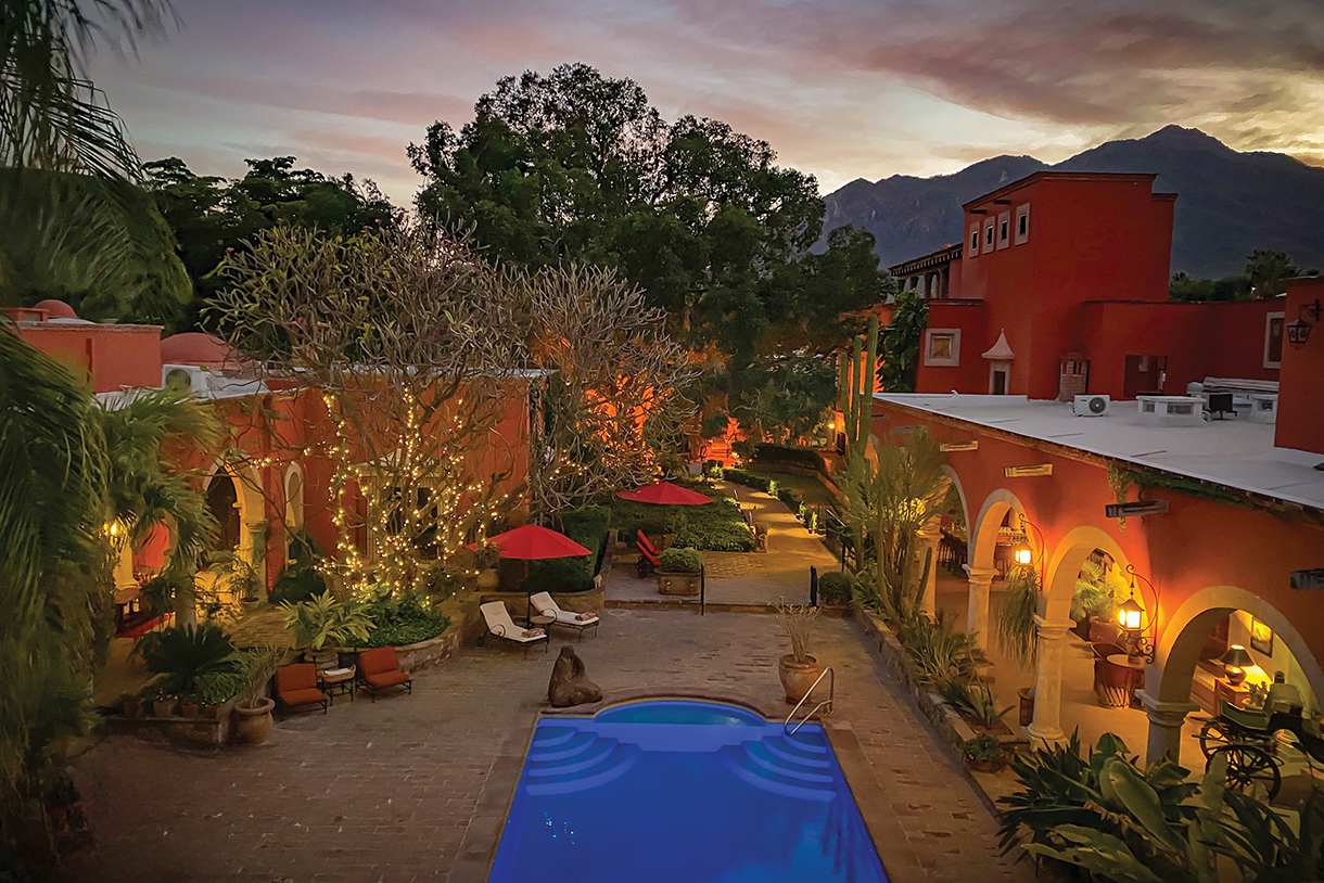 A night view of a lit courtyard at a hacienda with a swimming pool at the center. Lush green trees and buildings with arched hallways surround the courtyard. 