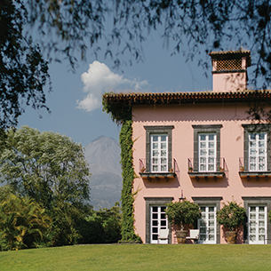 A large pink estate sitting on a lush green field with many trees. The building has many large windows on both floors.