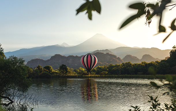 A blue, white, and red hot air balloon floating on top of a large calm lake. There are roling hills in the background.