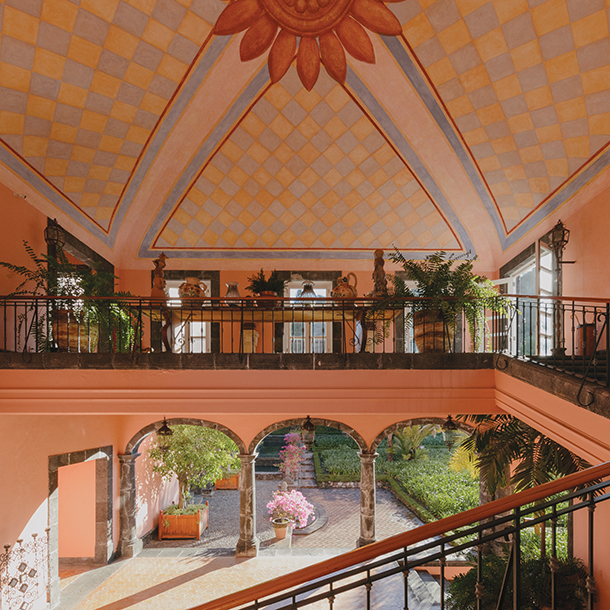 Interior of hacienda estate with balcony that overlooks the bottom floor. The high ceiling is colorfully painted with a floral motif.