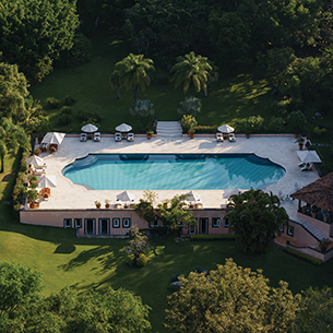 An aerial view of a large swimming pool surrounded by trees. There is a patio deck area next to the pool with lounge chairs and umbrellas. 