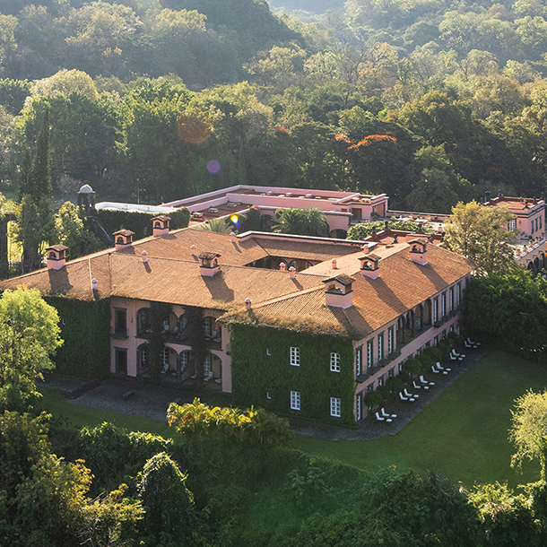 An aerial view of a large hacienda surrounded by lush, green highlands. The large estate has a red tile roof, many windows, and pink-colored stucco walls with some outer walls covered in greenery.)